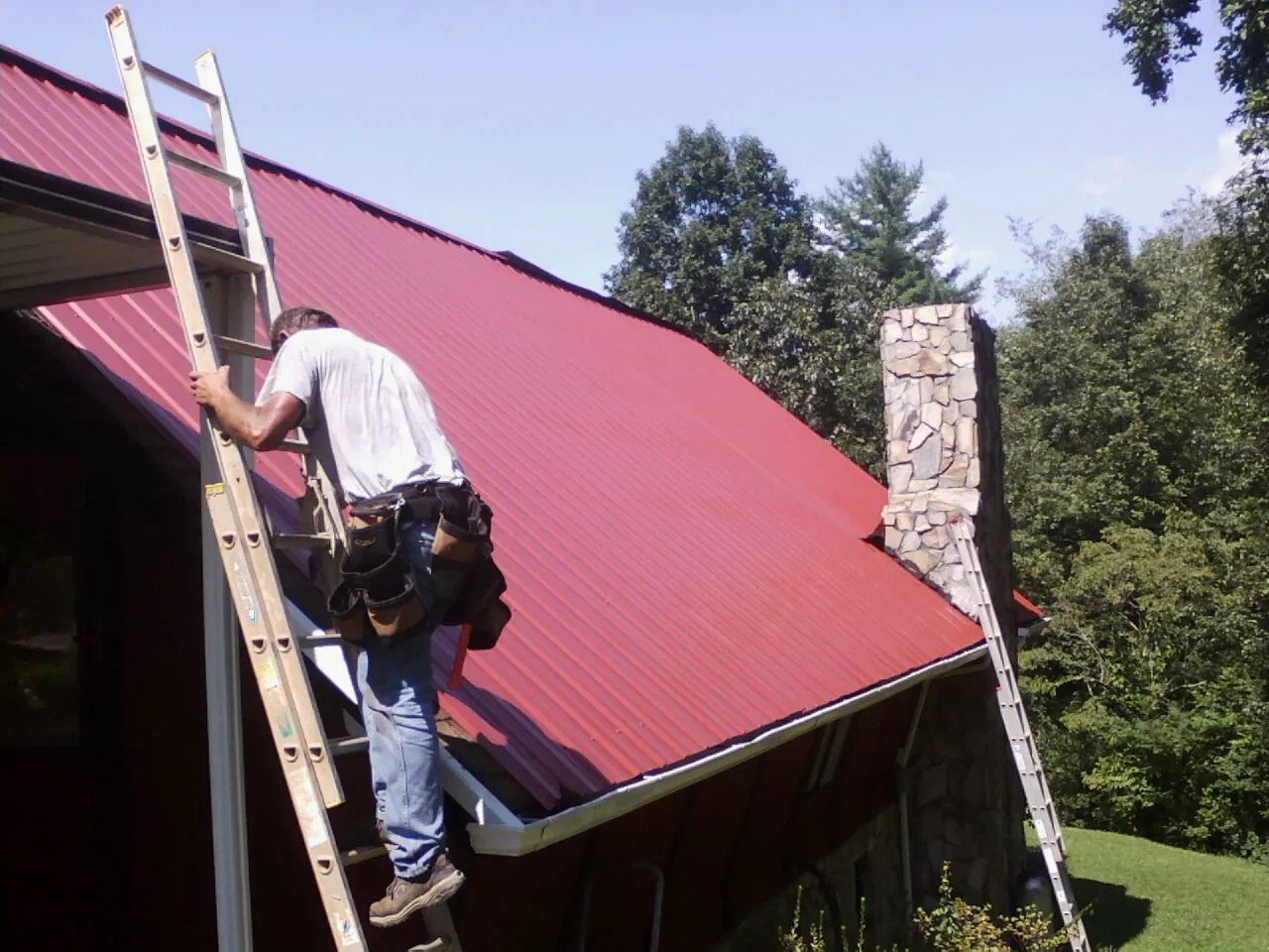 Roofer on a ladder beside a red metal roof and stone chimney — Soffit & Fascia Repair by Roofing Brothers in Harrogate